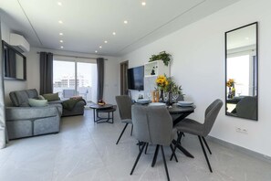 Ein modernes Wohnzimmer mit grauem Sofa, einem Esstisch mit Stühlen, einem Flachbildfernseher und einem großen Fenster mit Vorhängen.