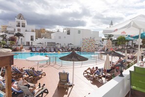 A pool area with lounge chairs and umbrellas, surrounded by buildings with balconies and a clear sky.