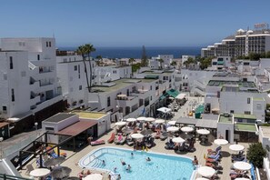 Un área de piscina en la azotea con varios niveles de edificios, una vista despejada del océano y una variedad de tumbonas y sombrillas.
