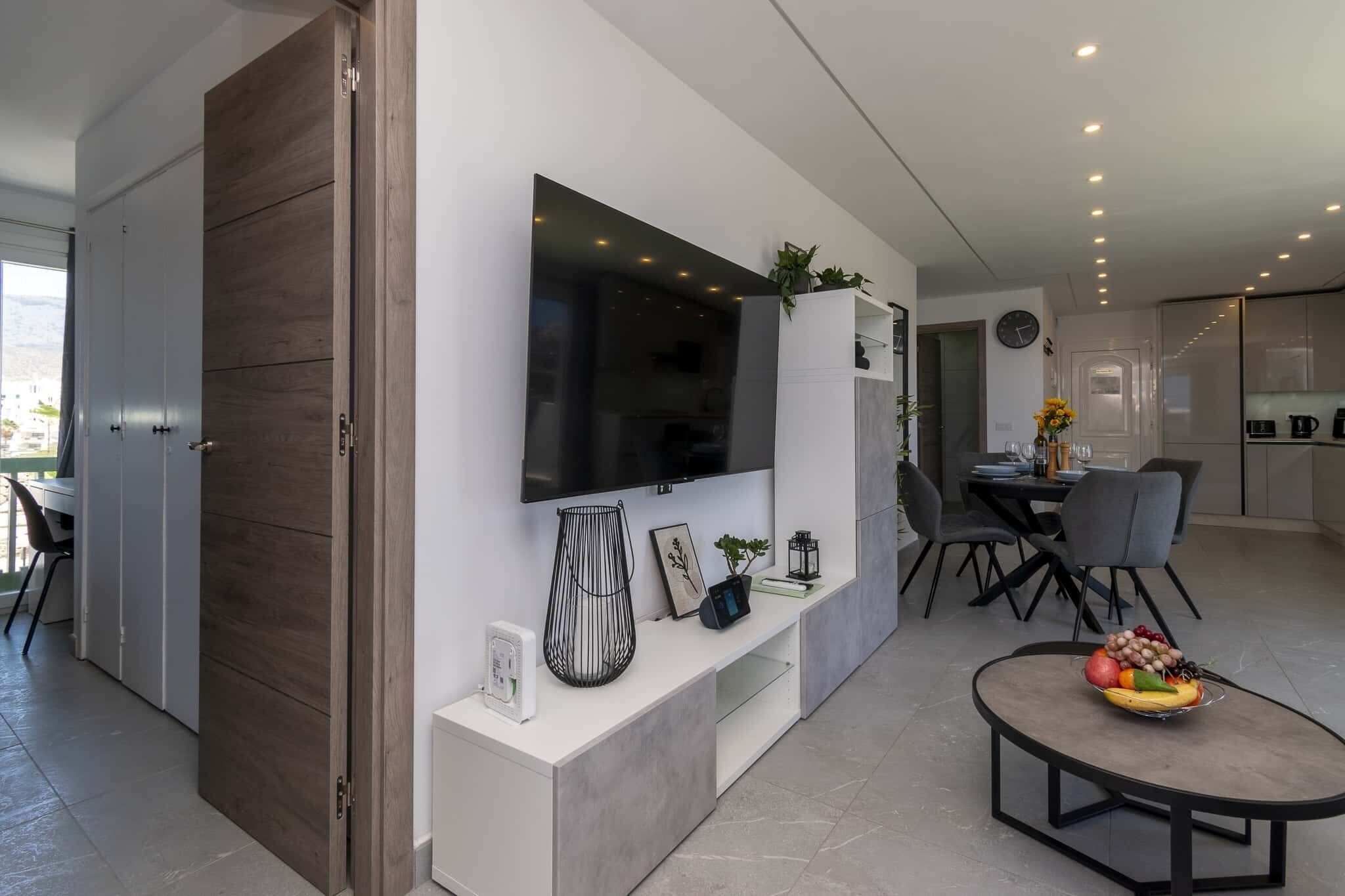 Ein modernes Wohnzimmer mit Flachbildfernseher, Essbereich mit Tisch und Stühlen sowie Küchenzeile mit Schränken und Geräten.