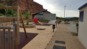 An outdoor playground with a wooden swing set, a red slide, and a yellow slide. There is a modern building with a unique geometric facade in the background. - MIRADORES DEL ISASA - VIURA-NEOSENS STUDIO APARTMENT (HERCE, La Rioja)