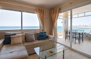 A modern living room with a beige sofa, glass coffee table, and a view of the ocean through large windows.