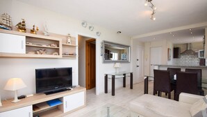 Ein modernes Wohnzimmer mit Flachbildfernseher, Glastisch und beiger Couch. Es gibt ein Holzregal mit Dekorationen und einen kleinen Tisch mit Lampe.