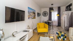 A modern living room with a flat-screen TV, a yellow armchair, a dining area with a table and chairs, and a patterned rug.