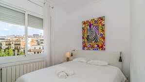 A hotel room with a large bed, a window with a view of buildings, a colorful artwork on the wall, and a nightstand with a lamp.