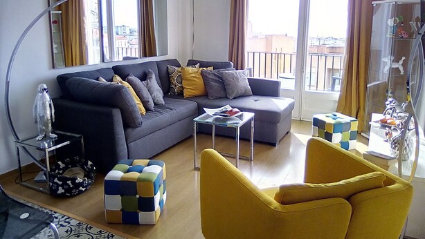 A modern living room with a grey sofa, yellow armchair, and a glass coffee table. There is a small dog bed and a colorful cube-shaped stool.