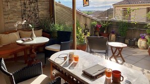 Un patio iluminado por el sol con una mesa de madera, una mesa redonda y un sofá. Hay plantas en macetas y vistas del barrio.