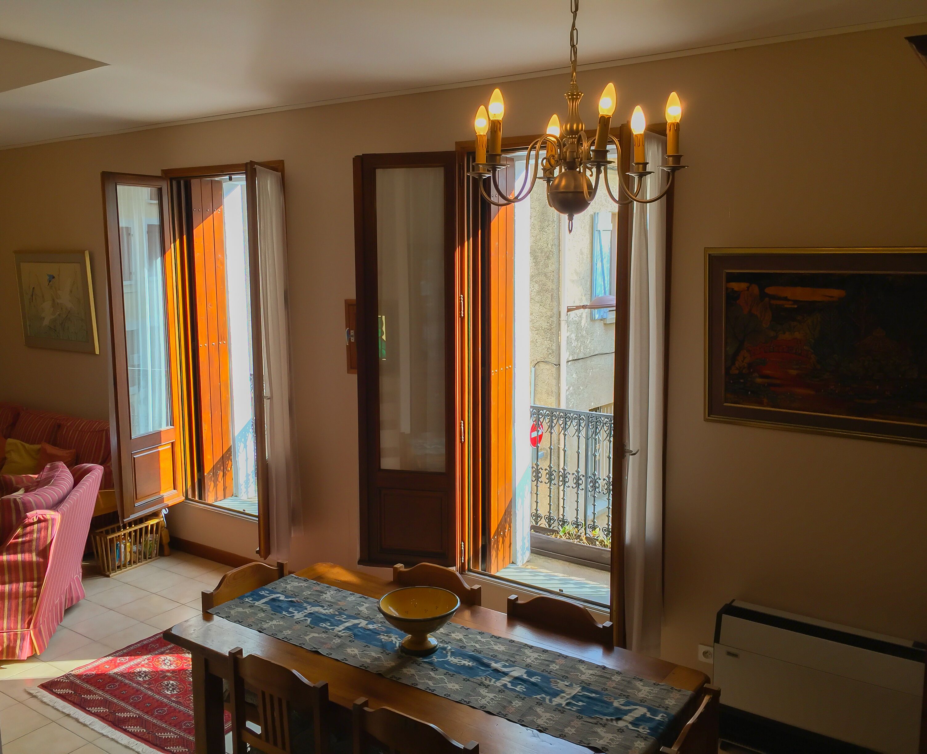 Un espace repas comprenant une table en bois, des chaises, un lustre et donnant sur le balcon extérieur.