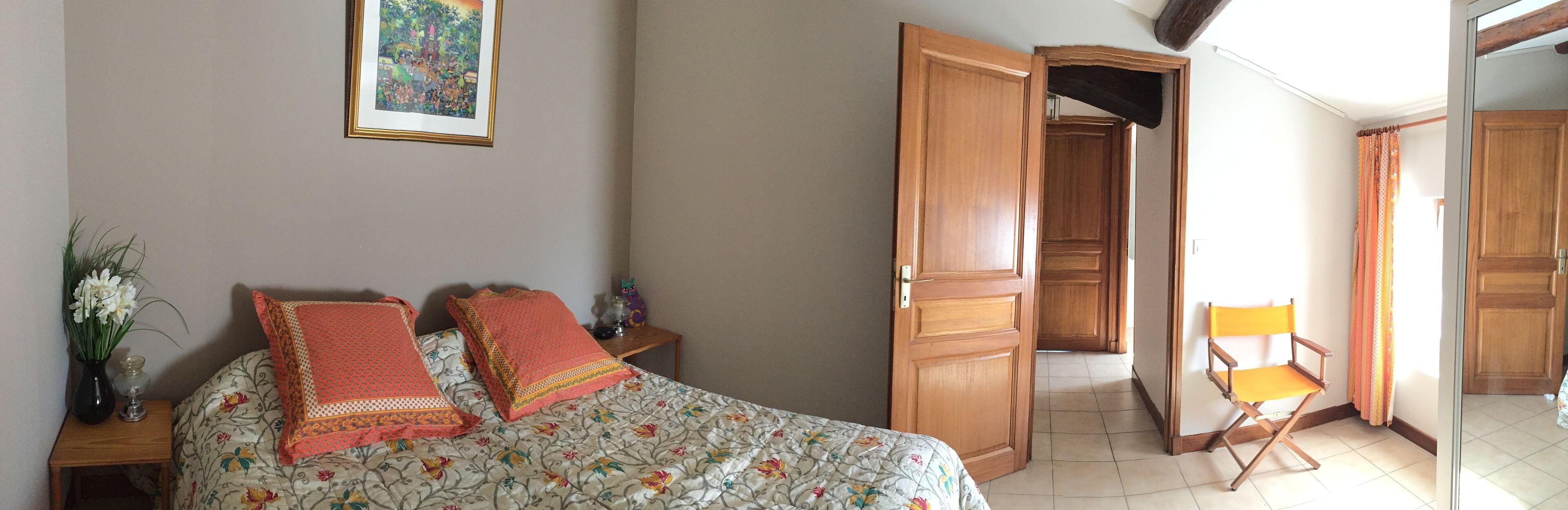 A bedroom with a bed, two pillows, a nightstand with a vase of flowers, a framed picture on the wall, and an open door leading to another room.