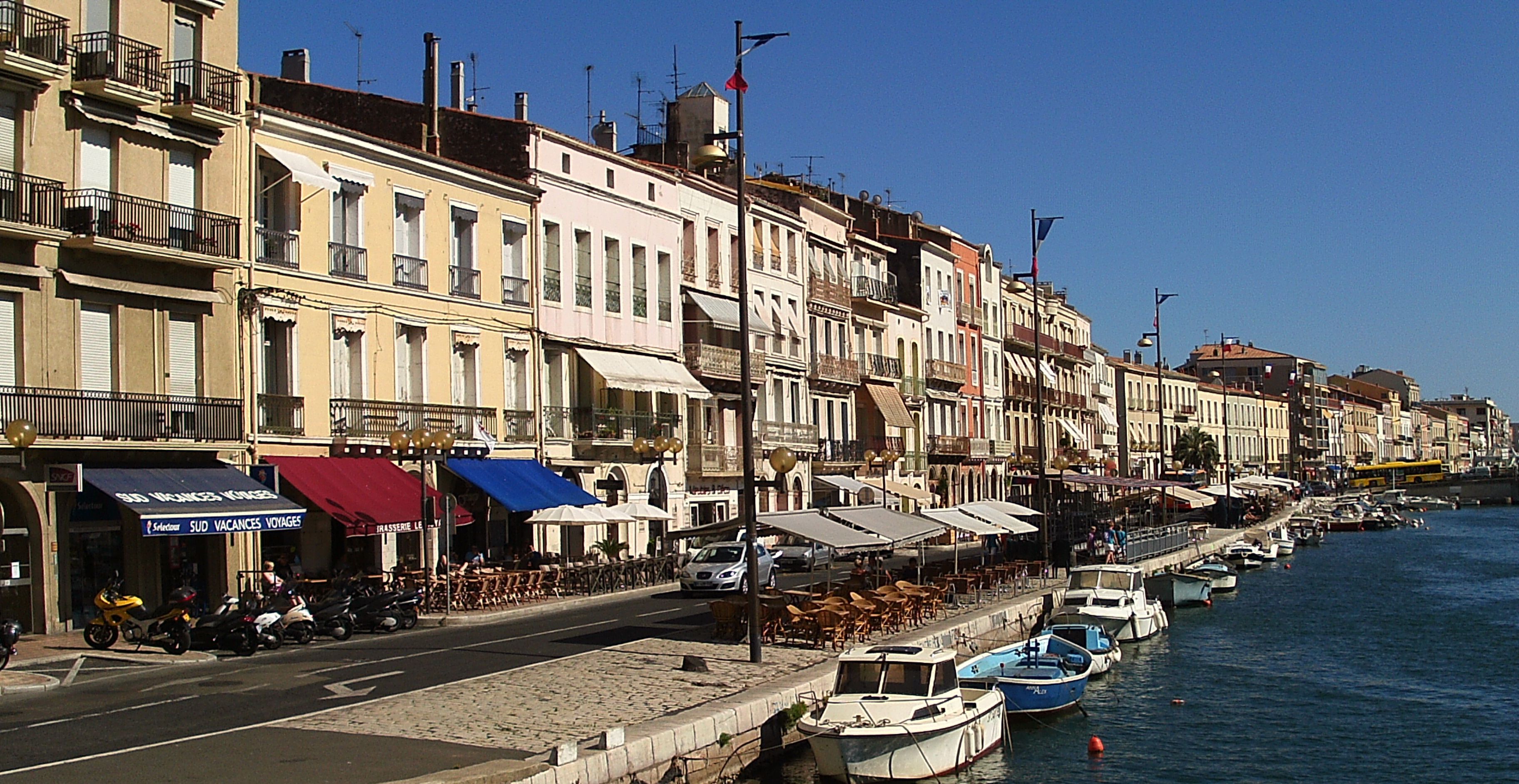 A waterfront street with buildings, outdoor seating, and boats docked along the water.