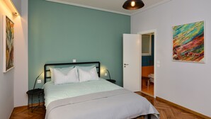 A bedroom with a bed, bedside tables, a nightstand, a door leading to another room, and a painting on the wall.