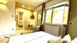 Une chambre d’hôtel avec un lit, une fenêtre, un radiateur, une table de chevet et une salle de bain visible à travers une porte ouverte.