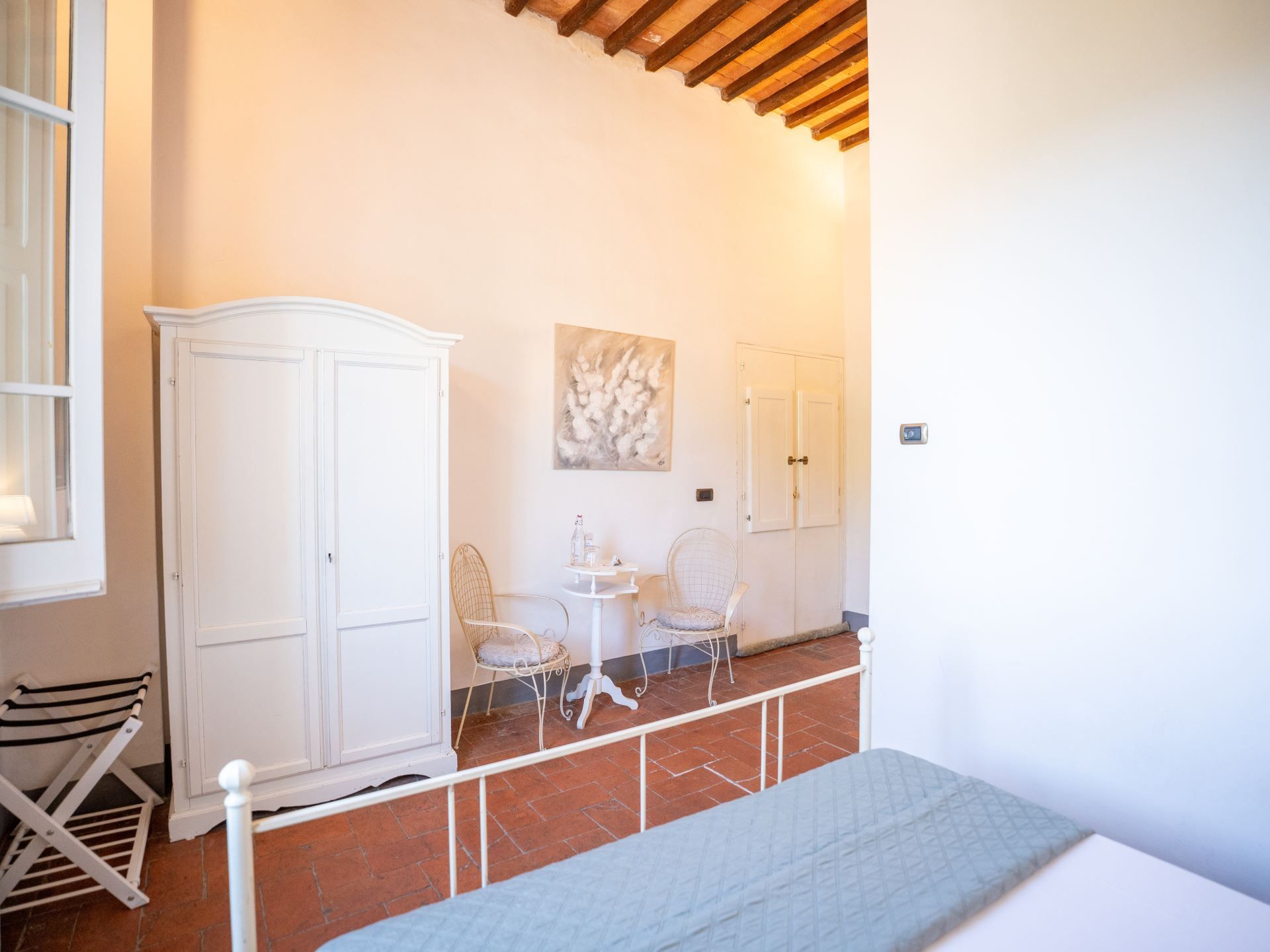 Een kamer met een bed, een witte kledingkast, twee stoelen, een klein tafeltje en een schilderij aan de muur.