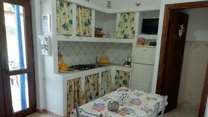 A compact kitchen with a gas stove, a sink, and a refrigerator. There is a dining table with a floral tablecloth and a chair. - Holiday home Kristin (Lipari)