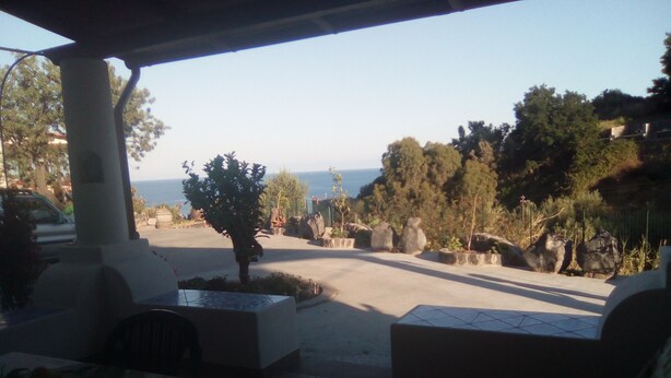 An outdoor seating area with a view of the sea, featuring a covered patio, stone planters, and a mix of trees and shrubs. - Holiday home Kristin (Lipari)