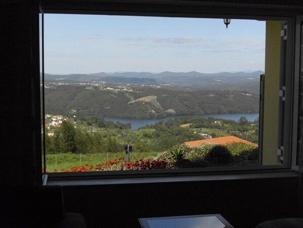 A room with a large window offering a view of a lake, hills, and a red-roofed building.
