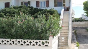 Un edificio de dos plantas con un balcón, entrada protegida por una reja y un pequeño jardín con arbustos y flores.