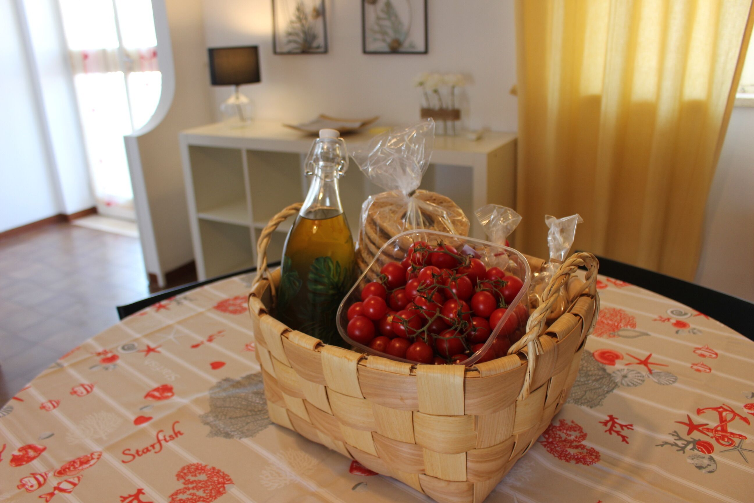 Een gevlochten mand op een tafel met een bloemen tafelkleed, met daarin een fles, een zak tomaten en een plastic zak.