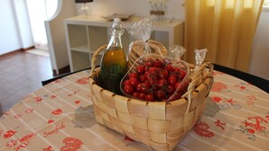 Una cesta tejida sobre una mesa con mantel floreado, que contiene una botella, un saco de tomates y una bolsa de plástico.