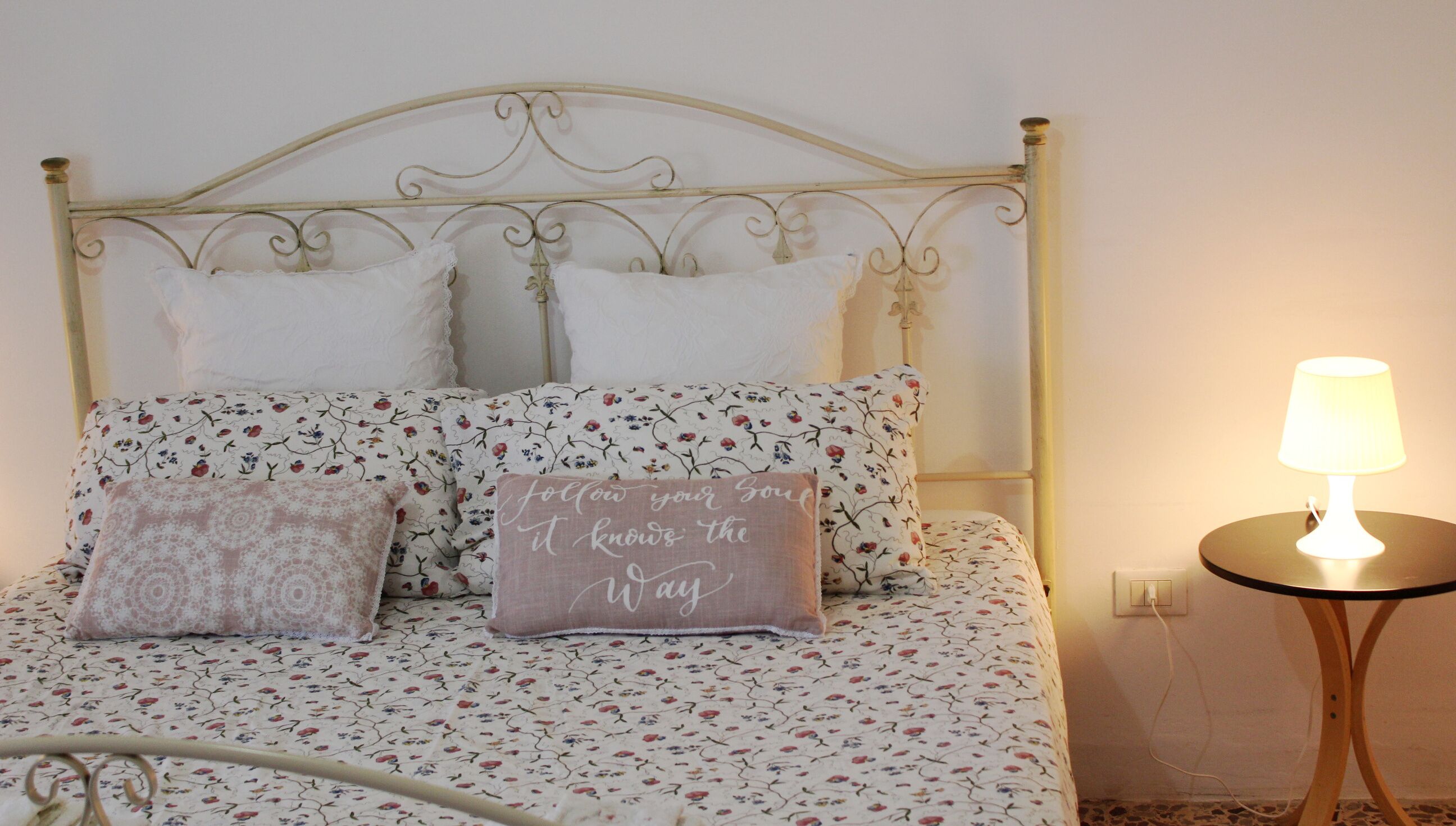 A neatly made bed with floral bedding and decorative pillows, a small bedside table with a lamp, and a wall socket.