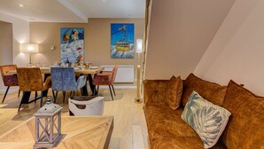 A modern living room with a wooden dining table, cushioned chairs, a brown sofa with a leaf-patterned pillow, and two large paintings on the walls.