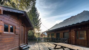 A rustic wooden cabin with a wooden deck and picnic table, surrounded by trees and mountains.