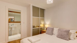 A hotel room with a bed, a bedside table with a lamp, a large window, and a bathroom visible through a glass door.