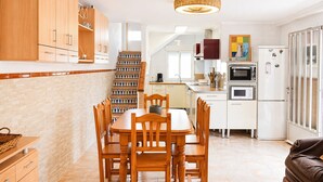 A modern kitchen with wooden cabinets, a dining table with chairs, a staircase with colorful steps, and a refrigerator.