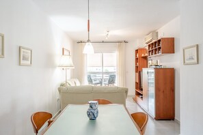 Un espace repas comprenant une table, des chaises, un vase, une suspension, un canapé, une fenêtre et un coin cuisine avec des placards.