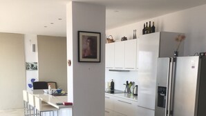 Una sala moderna con sofá, comedor y cocina con gabinetes blancos y un refrigerador de acero inoxidable.