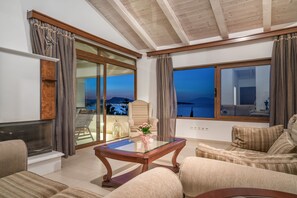 A spacious living room with a glass table, comfortable seating, a large window with a view, and a wooden ceiling.