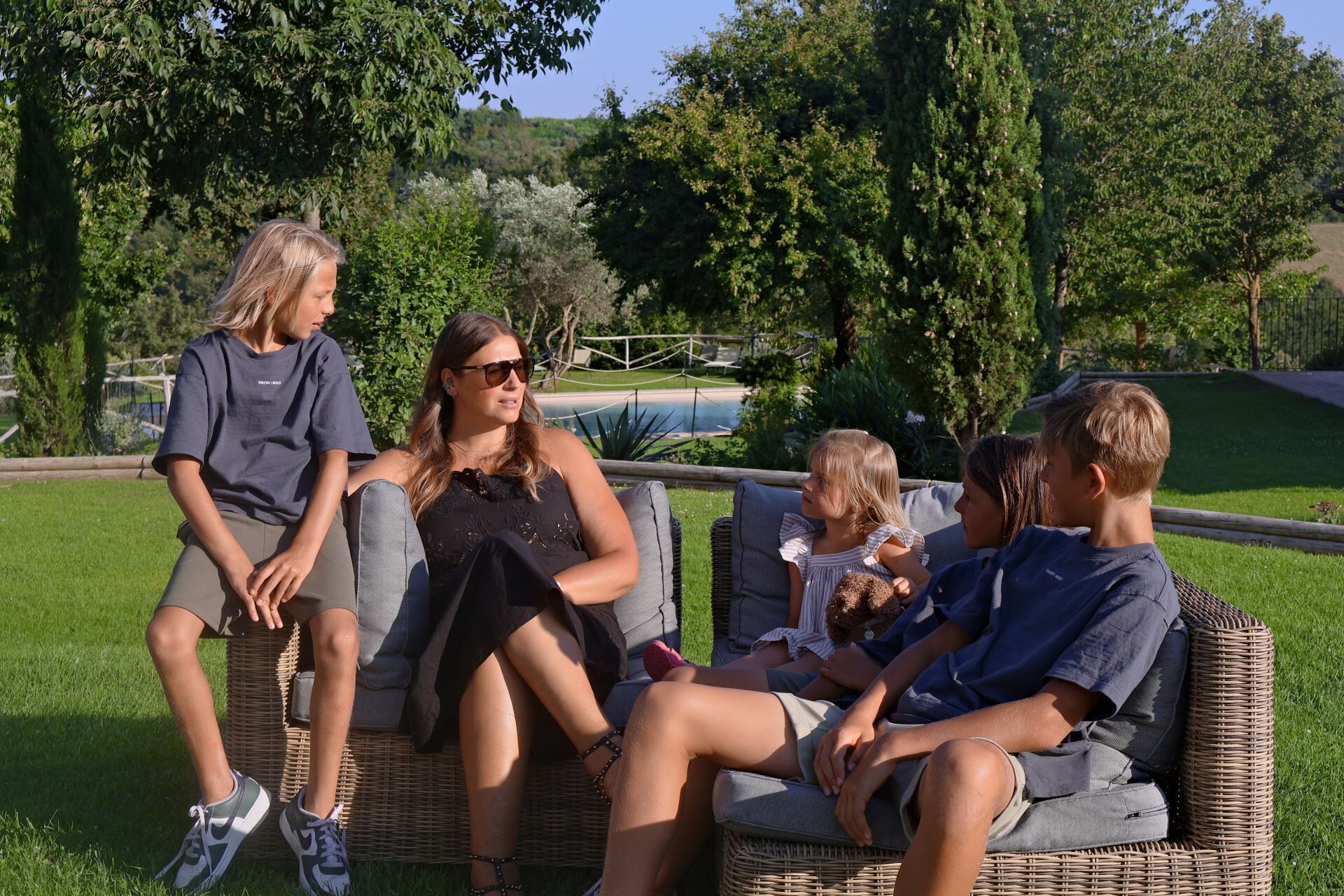Un grupo de personas sentadas en muebles de mimbre al aire libre, con una piscina y follaje en el fondo.