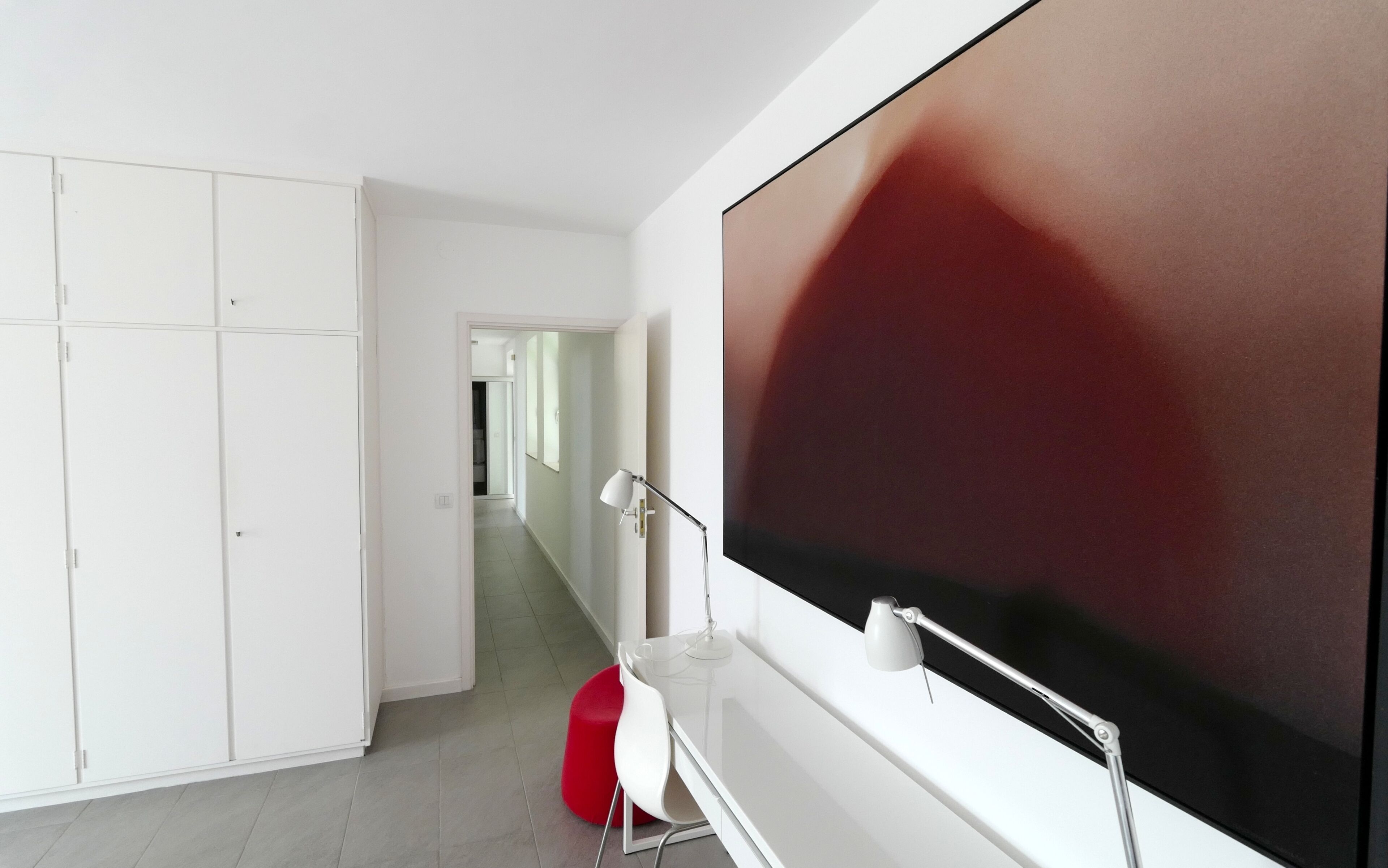 A modern room with a large flat-screen TV mounted on the wall, a white desk with a lamp, and a red chair.