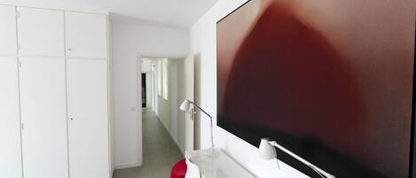 A modern room with a large flat-screen TV mounted on the wall, a white desk with a lamp, and a red chair.