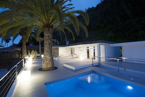 Un espace piscine moderne avec une piscine à l’eau d’un bleu limpide, des piliers en pierre et un coin repas équipé de tables et de chaises.