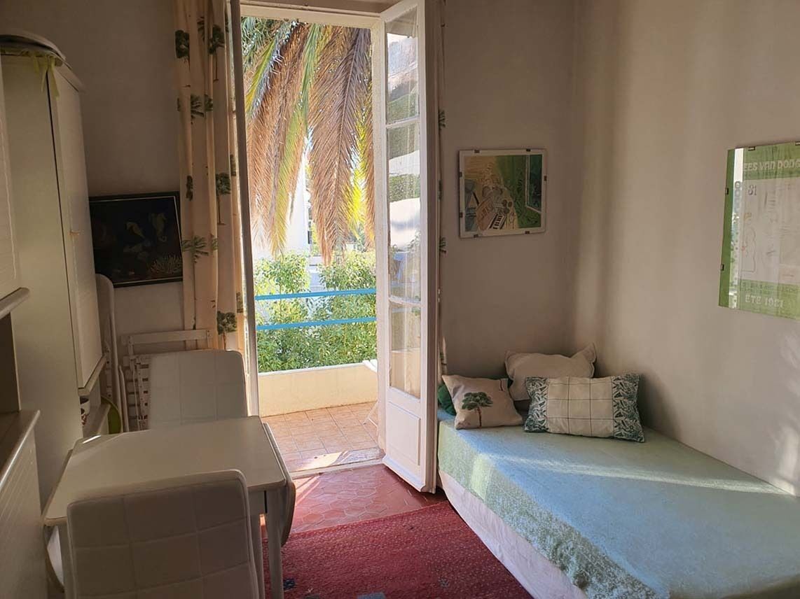 A room with a bed, a small table, a chair, a window with a view of greenery, and a door leading outside.
