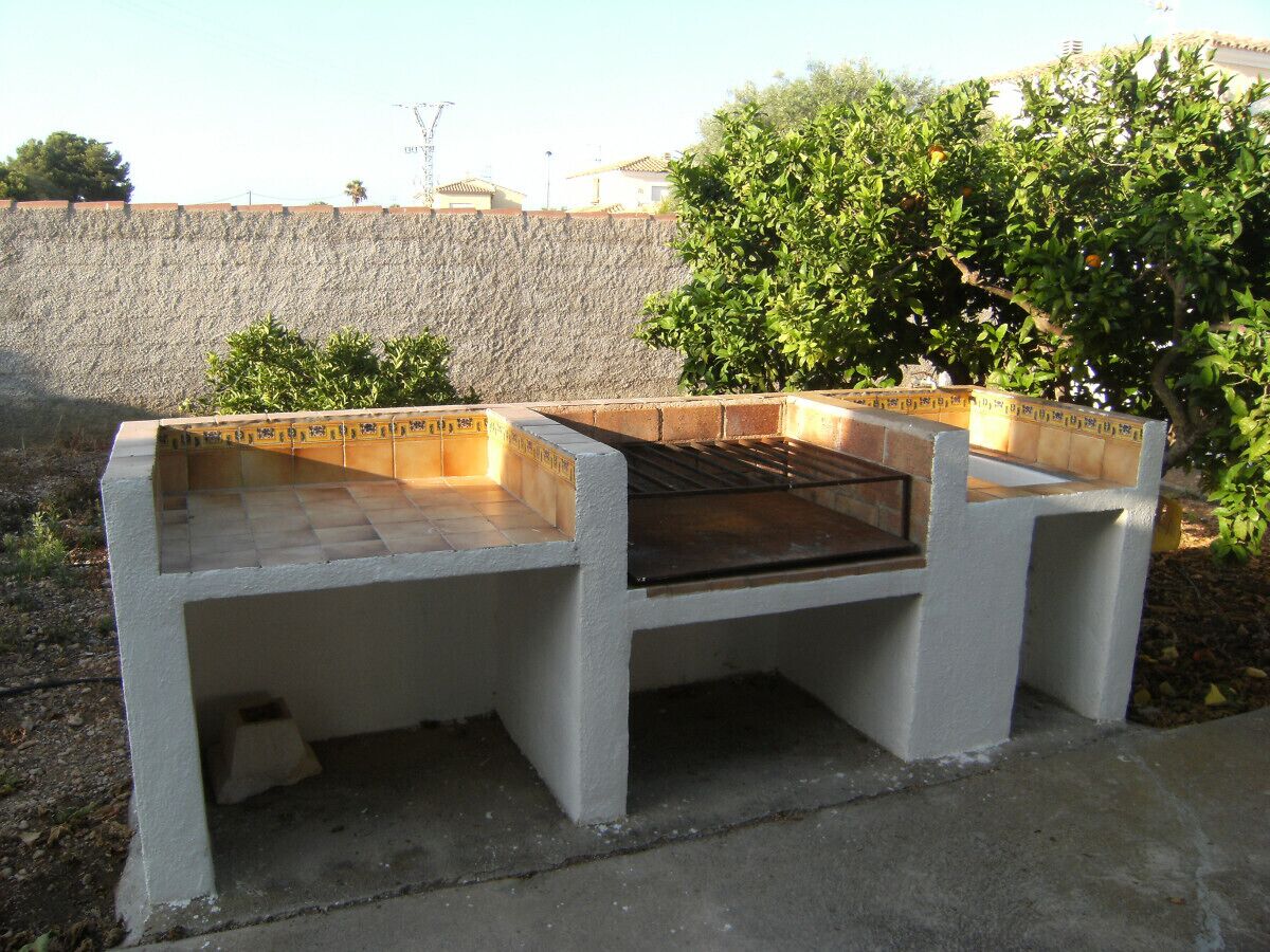 A concrete barbecue grill with a tiled top and a metal grate, surrounded by a stone wall and greenery.