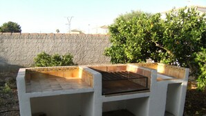 Una parrilla de concreto con cubierta de baldosas y rejilla metálica, rodeada de un muro de piedra y follaje.