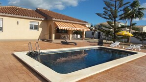 Una piscina rectangular con solárium empedrado, escalera de metal y zona de estar cubierta con toldo a rayas.