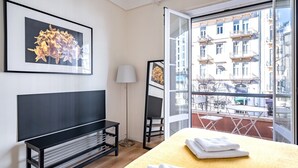 Een hotelkamer met een bed, een bankje, een vloerlamp en een groot raam met uitzicht op de stad.