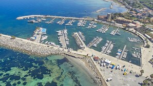 Una vista aerea di un porto turistico con numerose imbarcazioni ormeggiate, circondato da una cittadina costiera con edifici e un mare blu limpido.