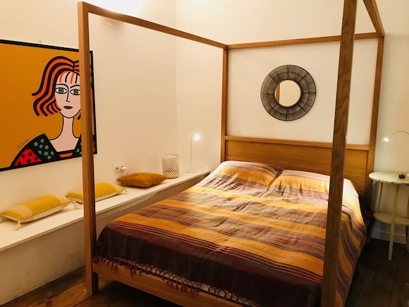 A four-poster bed with a striped bedspread, a round mirror on the wall, and a painting of a woman on the wall.