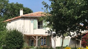 A two-story house with a tiled roof, a balcony, and a garden with sunflowers.