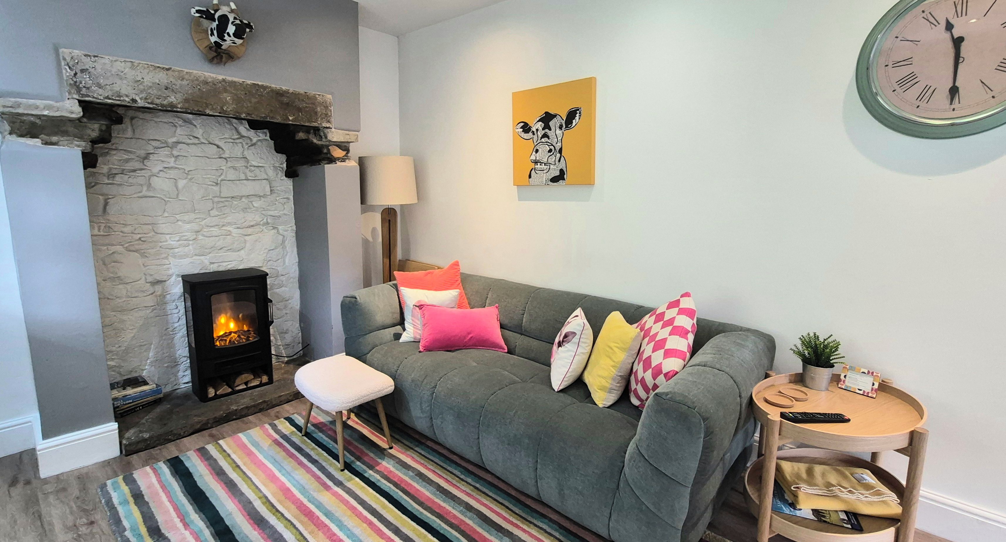 A cozy living room with a stone fireplace, a grey sofa with colorful cushions, a striped rug, and a round wooden side table with a remote control.