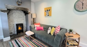 A cozy living room with a stone fireplace, a grey sofa with colorful cushions, a striped rug, and a round wooden side table with a remote control.