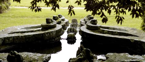 Une structure en pierre avec une fontaine centrale, entourée de pelouse et d’arbres.