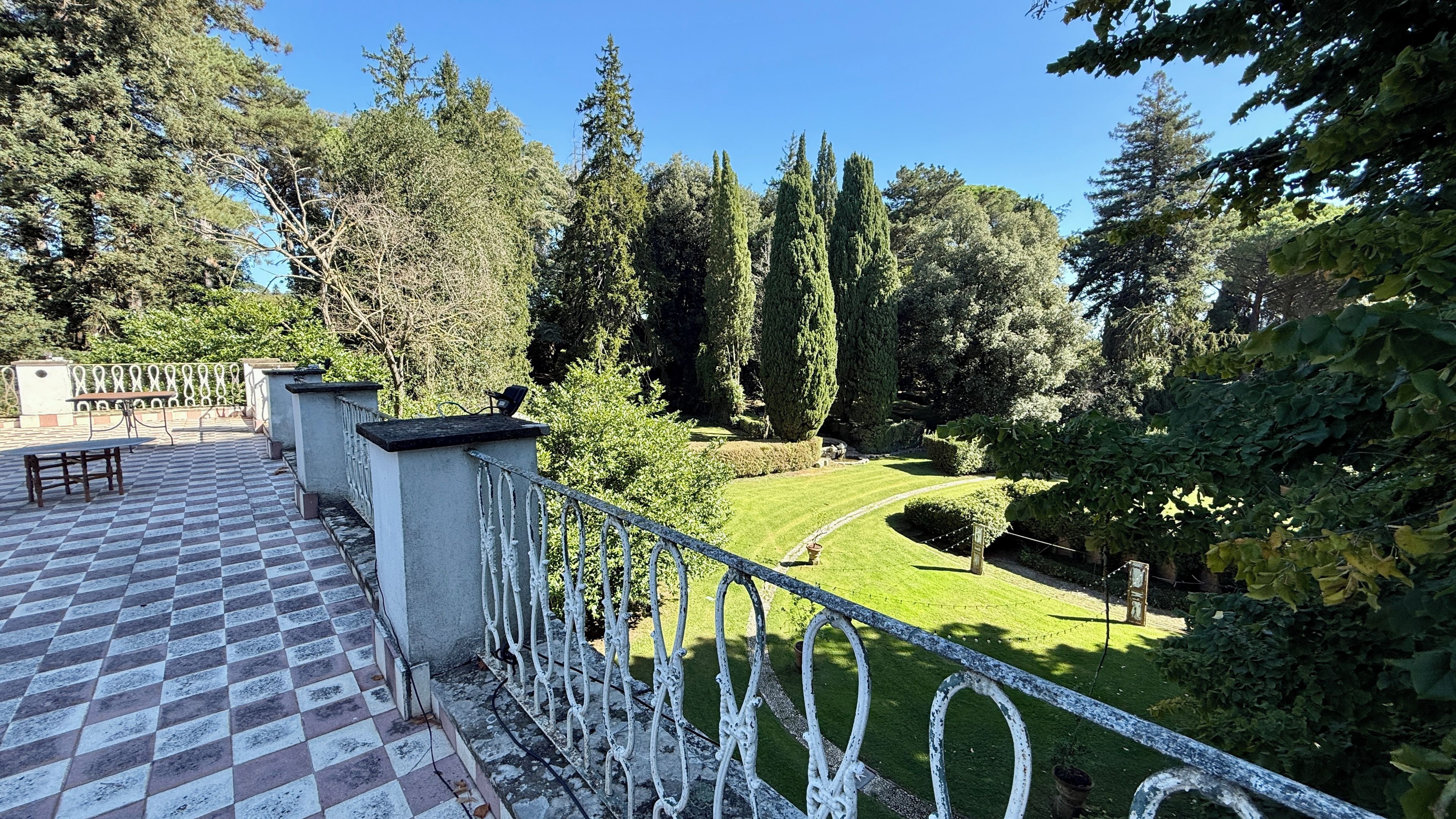 A terrace with a checkered floor, a metal railing, and a view of a well-maintained garden with trees and a lawn.