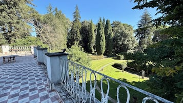 A terrace with a checkered floor, a metal railing, and a view of a well-maintained garden with trees and a lawn.