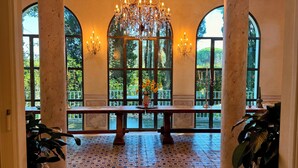 Un salón amplio con una gran mesa de madera, candelabros ornamentados y amplias ventanas de arco que ofrecen vistas de un follaje exuberante.