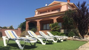 A two-story house with a tiled roof and a balcony. There are four lounge chairs with striped cushions on a green lawn in front of the house. - Villa Mercè. Golden Coast (Botarell)